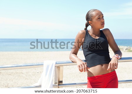 Athletic african american young woman relaxing with a towel, leaning on a shiny railing exercising by the seaside during a sunny summer day. Health and sport lifestyle and wellness outdoors.
