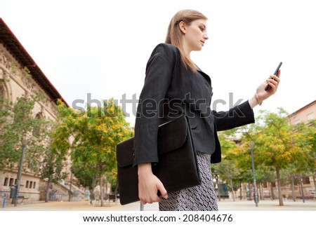 Side portrait of an elegant young professional business woman standing by a grand stone building in the city smiling, holding and using a smartphone. Business people with technology, outdoors.