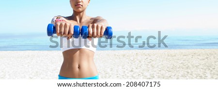 Portrait of a fit beautiful teenager girl exercising on a white sand beach, lifting small weights during an exercise and sport routine against an intense blue sky. Sporty healthy lifestyle outdoors.