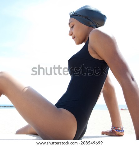 Side view of a healthy sporty african american young woman swimmer sitting on a beach with goggles and swimming cap, being thoughtful and focused against a sunny blue sky. Sport lifestyle outdoors.