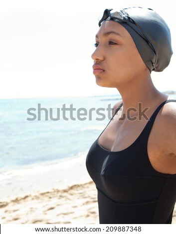 Side close up portrait of a healthy african american young woman swimmer on a beach with goggles and a swimming cap, training outdoors feeling strong and confident. Sport lifestyle outdoors.