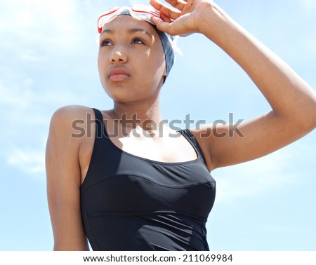 Portrait view of a fit, healthy young black woman swimmer sitting on a rock by the sea against a sunny blue sky, holding swimming goggles and looking thoughtful. Sport lifestyle, outdoors.