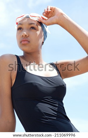 Portrait view of a fit, healthy young black woman swimmer sitting by the sea against a sunny blue sky, holding swimming goggles and looking thoughtful. Sport lifestyle, outdoors.