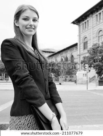 Black and white portrait of a smiling professional business woman in a classic city with stone buildings, smiling and holding a briefcase folder under her arms. Business people at work, outdoors.