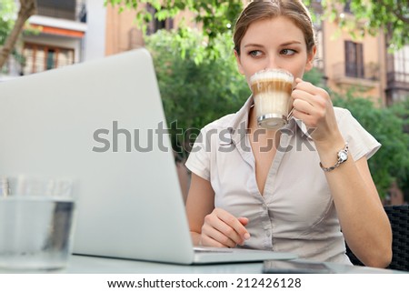 Attractive professional young business woman sitting at a coffee shop drinking a beverage and using laptop computer and smartphone technology in the financial city, outdoors. Business people at work.