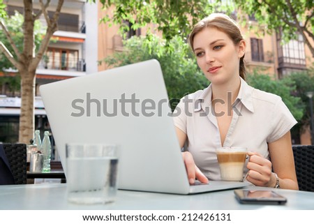 Attractive professional young business woman sitting at a coffee shop smiling, drinking a beverage and using laptop computer and smartphone technology in the financial city, outdoors.