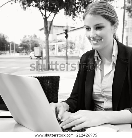 Black and white portrait of an attractive young professional business woman sitting at a cafe terrace drinking coffee, using a laptop computer in a city, outdoors. Smiling businesswoman.