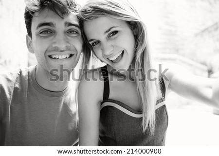 Black and white portrait of a young attractive tourist couple using a smartphone to take a selfie picture of themselves on holiday while visiting a touristic destination city, having fun outdoors.