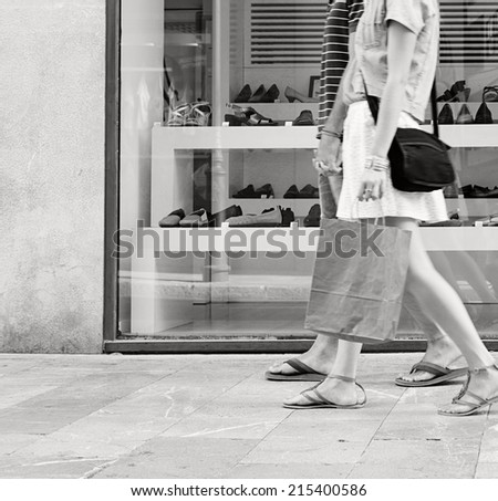 Side black and white view of a young couple lower body section walking in shopping street with a shoe store window display, shopping bags and spending money on holiday. Consumer lifestyle, outdoors.