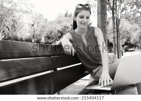 Black and white view of an attractive business woman sitting and working on her laptop computer while relaxing on a bench in a city street with trees during a sunny day, outdoors.