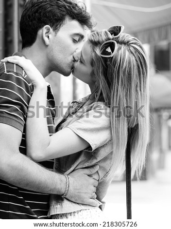 Black and white side portrait view of a young and attractive couple on holiday, kissing and embracing while shopping, traveling in a destination city street, outdoors. Love, passion and relationships.