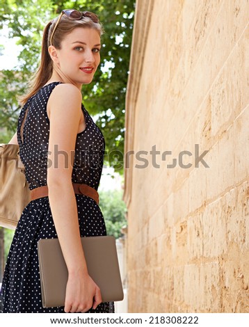 Rear view of a professional young businesswoman walking by an old stone building wall while commuting to work in a classic city street, turning and smiling at the camera, outdoors.