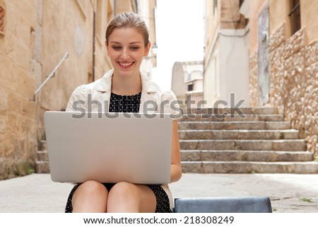 Attractive smiling young professional business woman sitting on the steps of an old stone street using a laptop computer working outdoors. Connectivity and wireless internet browsing.