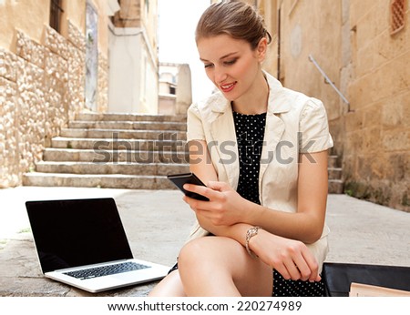 Attractive young business woman using smartphone and laptop computer while sitting on a stone steps street in a classic city and smiling outdoors. Professional corporate people and technology.