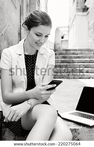 Black and white portrait of attractive young business woman using smartphone and laptop computer while sitting on a stone steps in a classic city, smiling outdoors. Professional people and technology.