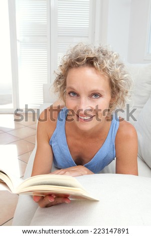 Beauty portrait of an attractive middle aged healthy woman relaxing on a white sofa at home, laying down and reading a book having a break, smiling. Interior home living and well being lifestyle.