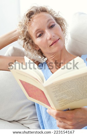Portrait of a beautiful middle aged woman laying down and relaxing on a white sofa at home, holding and reading a book, smiling having a break indoors. Home living and relaxing lifestyle interior.