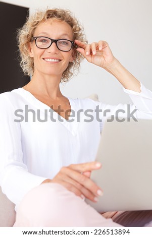 Portrait of a confident mature professional woman sitting on a white sofa at home using a laptop computer to work, indoors. Aspirational businesswoman wearing glasses and working from home.