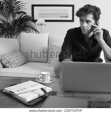 Black and white portrait of a professional businessman sitting on a coach at home, working on his laptop computer and making a phone call with a smartphone, indoors. Working from home office.
