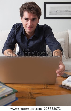 Attractive young professional business man sitting on a white coach in his home living room smiling, working on his laptop computer and looking at paperwork, indoors. Working from home office.