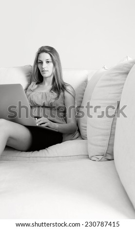 Black and white view of attractive professional business woman using laptop computer to browse the internet, sitting on a space white wall living room with a sofa, home interior. Space for type.