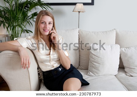 Attractive young professional business woman sitting on a white coach at home living room smiling, having a telephone conversation on her smartphone device, indoors. Working from home office.