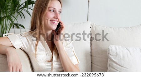 Panoramic view of an attractive professional businesswoman sitting on a coach at home living room, making a phone call with a smartphone, joyfully smiling indoors. Working from home office.