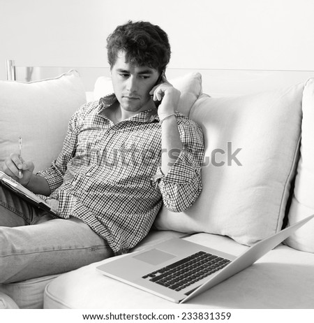 Black and white portrait of business man sitting on sofa in home living room, making a call on his smartphone and using a laptop computer. Professional businessman working from home, sitting on sofa.