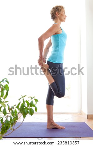 Side view of a mature healthy sporty woman using a yoga mat to exercise and stretch her body in a light and airy interior. Fit professional woman exercising and stretching her body, indoors.