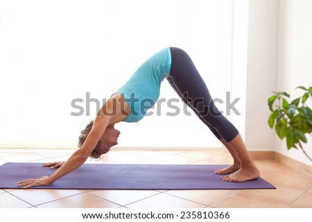Side view of a mature healthy sporty woman using a yoga mat to exercise and stretch her body in a light and airy interior. Fit professional woman exercising and stretching her body, indoors.