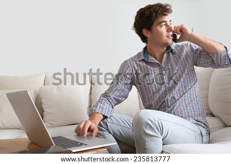 Working business man having a phone conversation on a smart phone and using a laptop working from home office. Young professional man using technology in a home living room, interior.