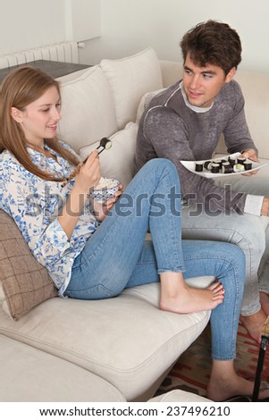Attractive young couple enjoying eating Japanese sushi and maki food at home, sitting on white couch in a home living room, sharing food and having a good time together. Man offering dish fresh food.