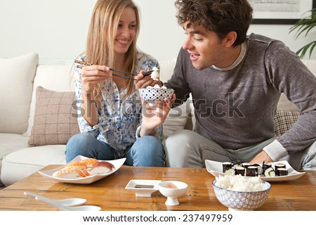 Attractive young couple enjoying eating Japanese sushi and maki food at home, sitting on a white couch in a home living room, sharing food and having a good time, smiling together. Eating fresh food.