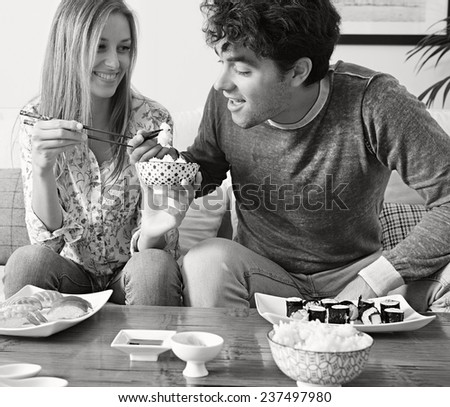 Black and white portrait of attractive couple eating Japanese sushi and maki food at home, sitting on a white couch, sharing food and having a good time, smiling together. Eating fresh food.