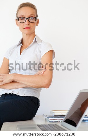 Middle aged senior office manager sitting and relaxing on her desk at work, confident and strong with her arms crossed, wearing reading glasses. Professional businesswoman in office, interior.