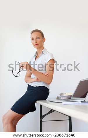 Mature senior office manager sitting and relaxing on her desk at work, confident and strong, holding reading glasses in her hand. Professional businesswoman in office interior. Technology in business.