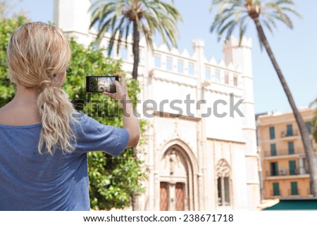 Rear portrait view of a young tourist girl holding up a smartphone device to point and take pictures of a monument while in a destination city on holiday. Vacation travel and technology networking.