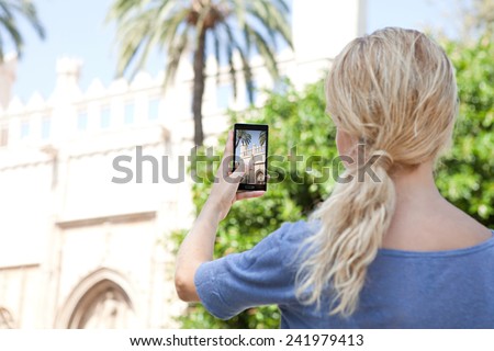 Rear portrait view of a young tourist girl holding up a smartphone device to take pictures of a monument while visiting a destination city on holiday. Vacation travel and technology networking.