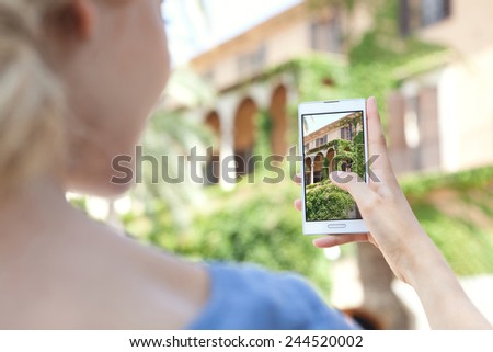 Rear portrait view of young tourist girl holding up a smartphone device to point and take pictures of monument while visiting a destination city on holiday. Vacation travel and technology networking.