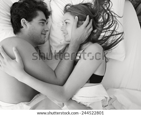 Overhead black and white portrait of an attractive young couple caressing laying in bed together in lingerie being romantic. Couple in a relationship caressing in a white bed, home interior lifestyle.