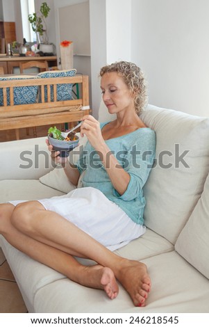 Attractive mature woman sitting and relaxing on a white couch at home eating a small green salad, home interior. Senior woman eating healthy food. Wellness and well being indoors. Lifestyle.