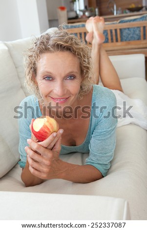 Attractive and mature healthy woman eating a red apple at home while relaxing on a white sofa in a home living room kitchen and smiling indoors. Healthy eating and well being lifestyle, interior.