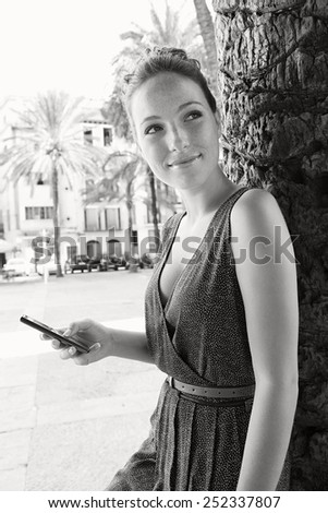 Black and white portrait of a young professional tourist woman visiting a picturesque square in a destination city with palm trees, using smartphone mobile on holiday. Travel and technology lifestyle.