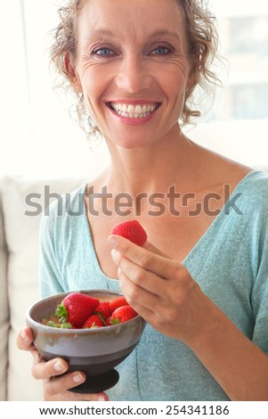 Close up portrait of a mature woman joyfully smiling and eating red strawberries at home relaxing on a white sofa in a home living room, indoors. Healthy eating and well being lifestyle, interior.
