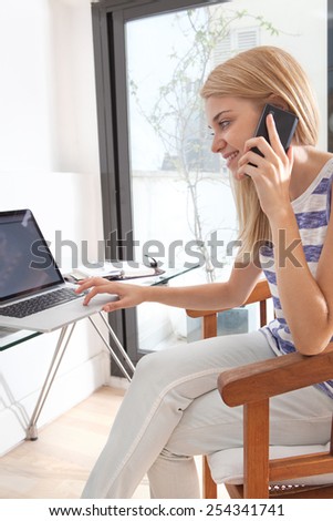 Portrait of a young woman working at her home office space with smartphone mobile having a call conversation. Student using a laptop computer at her home work desk. Lifestyle and technology, interior.