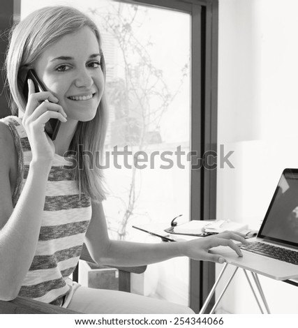 Black and white portrait of teenager girl college student at her work desk at home, using smartphone technology and a laptop computer to work and study. Professional woman with mobile phone indoors.