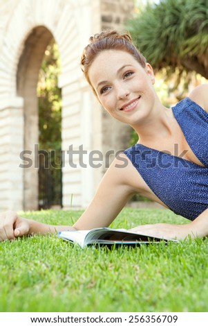 Attractive joyful tourist woman relaxing in a monument\'s park grass reading a travel guide book visiting a destination city on a summer holiday. Smart girl outdoors. Travel and vacation lifestyle.