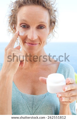 Close up beauty portrait of a mature attractive woman in her holiday resort hotel balcony, with sea views and blue sky, applying face cream on her skin, outdoors. Beauty and lifestyle and well being.