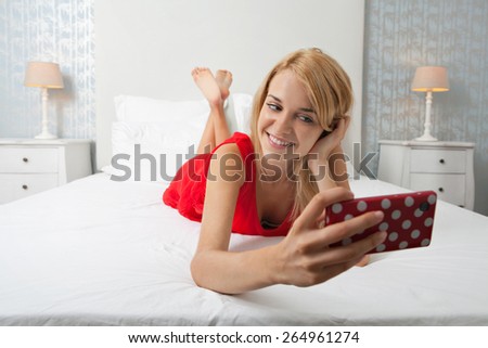 Portrait of attractive teenager woman laying in a stylish home bedroom using a smartphone device to take selfies pictures of herself, networking on line. Home interior technology lifestyle, indoors.