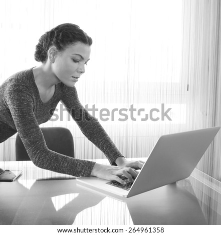 Black and white portrait of professional business woman at her office space work desk typing on laptop computer against a bright window with reflections on glass table. Business finance, interior.